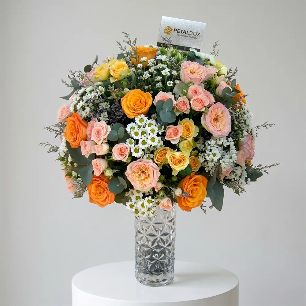 Elegant orange and blush rose vase arrangement featuring premium peach garden roses, orange roses, delicate spray roses, white daisy blooms, eucalyptus foliage, and seasonal fillers styled in a luxury crystal glass vase.